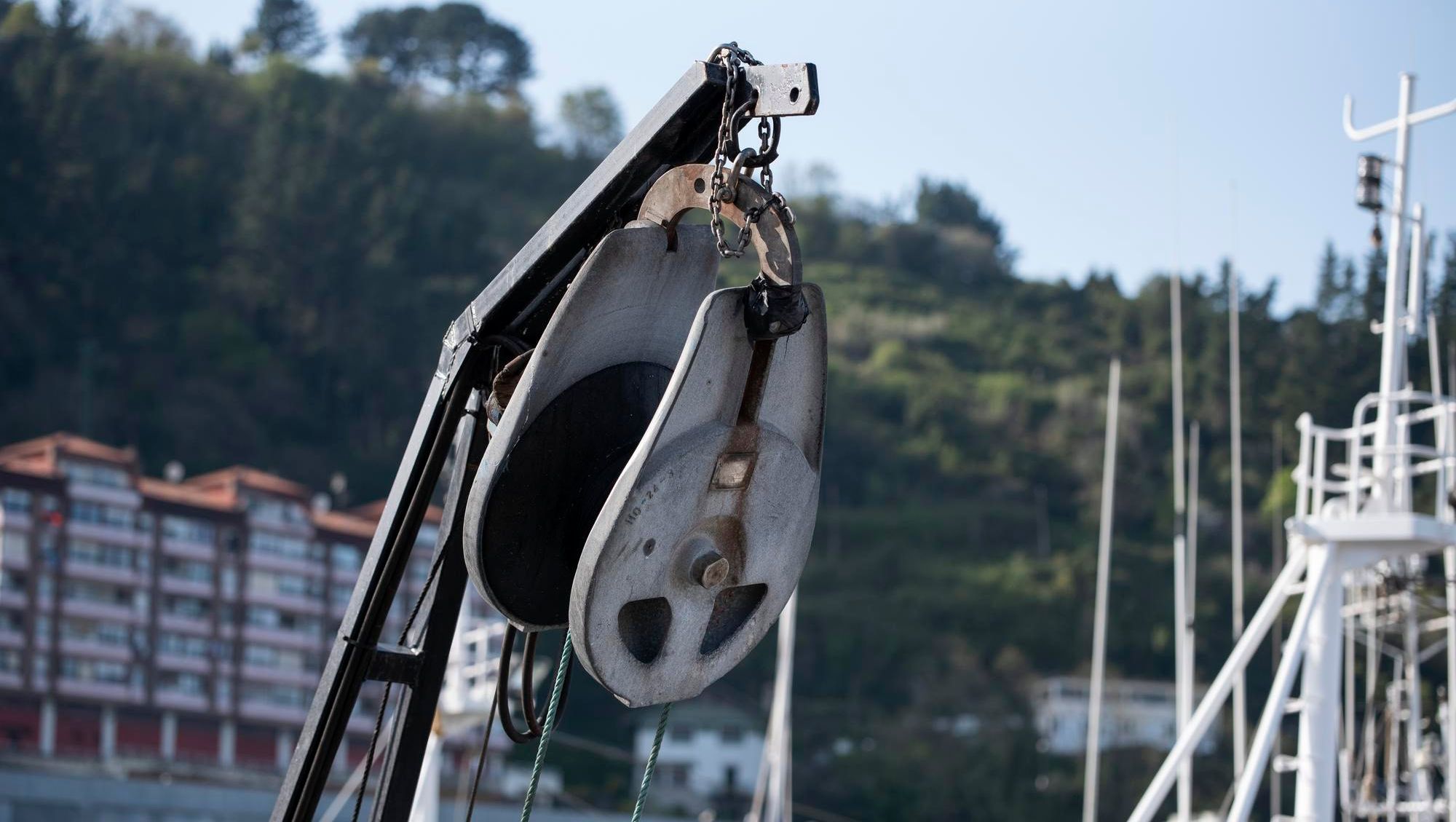 Nærbilde av blokk og talje på båt med mast og bebyggelse i bakgrunn.